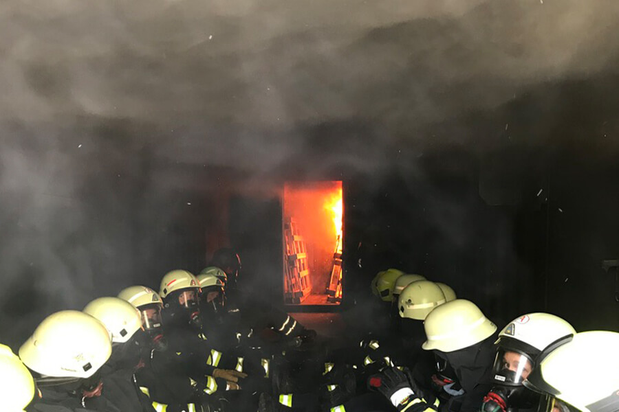 Feuerwehrkameraden sitzend im Rauch mit Atemschutzmasken, im Hintergrund schlagen Flammen durch eine Tür